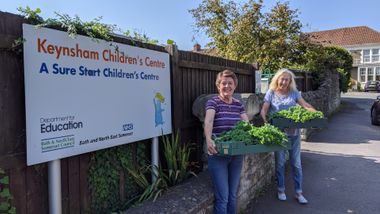 Two people holding a box of kale, stood outside next to the sign for Keynsham Children's Centre