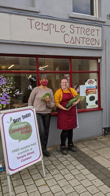 Two people standing outside Temple Street Canteen and the Keynsham Community Fridge in Keynsham posing with donated vegetables including a large marrow.